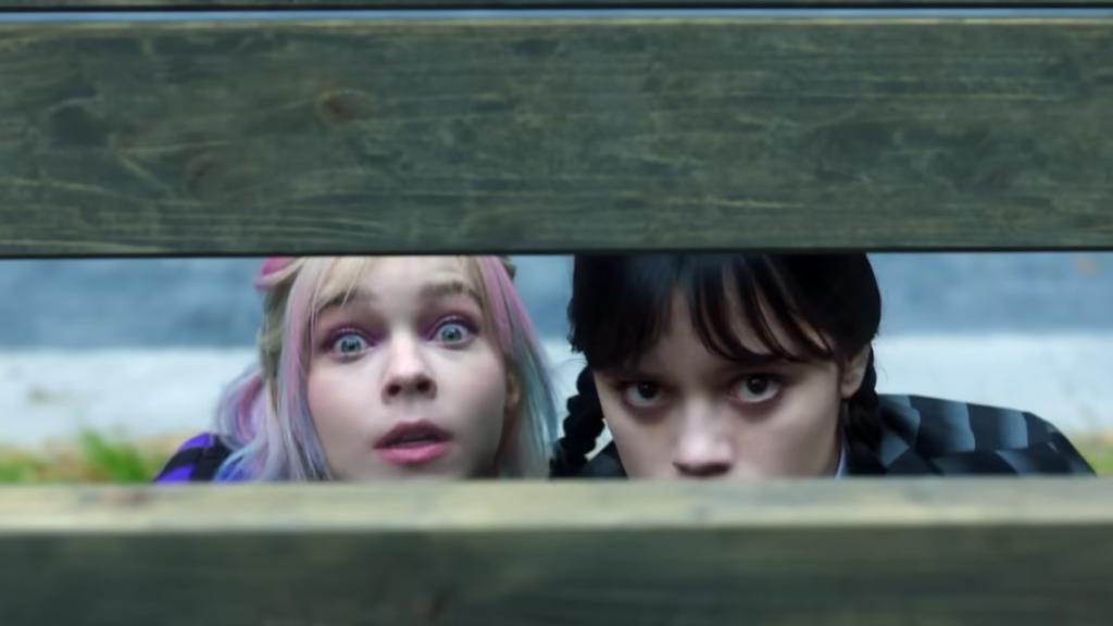 Enid and Wednesday looking through a hole in a fence