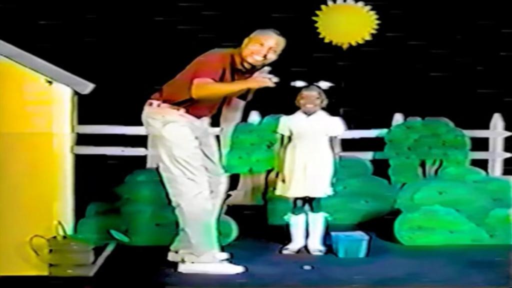 Live action shot of Sam Colton standing next to Rebecca, smiling at pointing at the camera